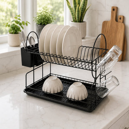 A black wire double-dish drying rack on a white marble kitchen counter, holding white plates, bowls, and clear drinking glasses, with plants and wooden cutting boards in the background.