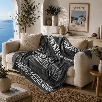 A dark gray throw blanket with a white Greek key pattern is draped over a beige sofa in a bright, modern living room with a view of the blue sea.